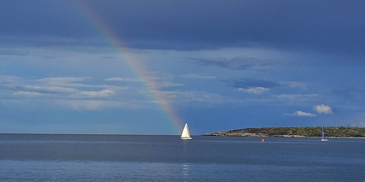 A faint rainbow descends through blue storm clouds toward a white sailboat on calm sea.
